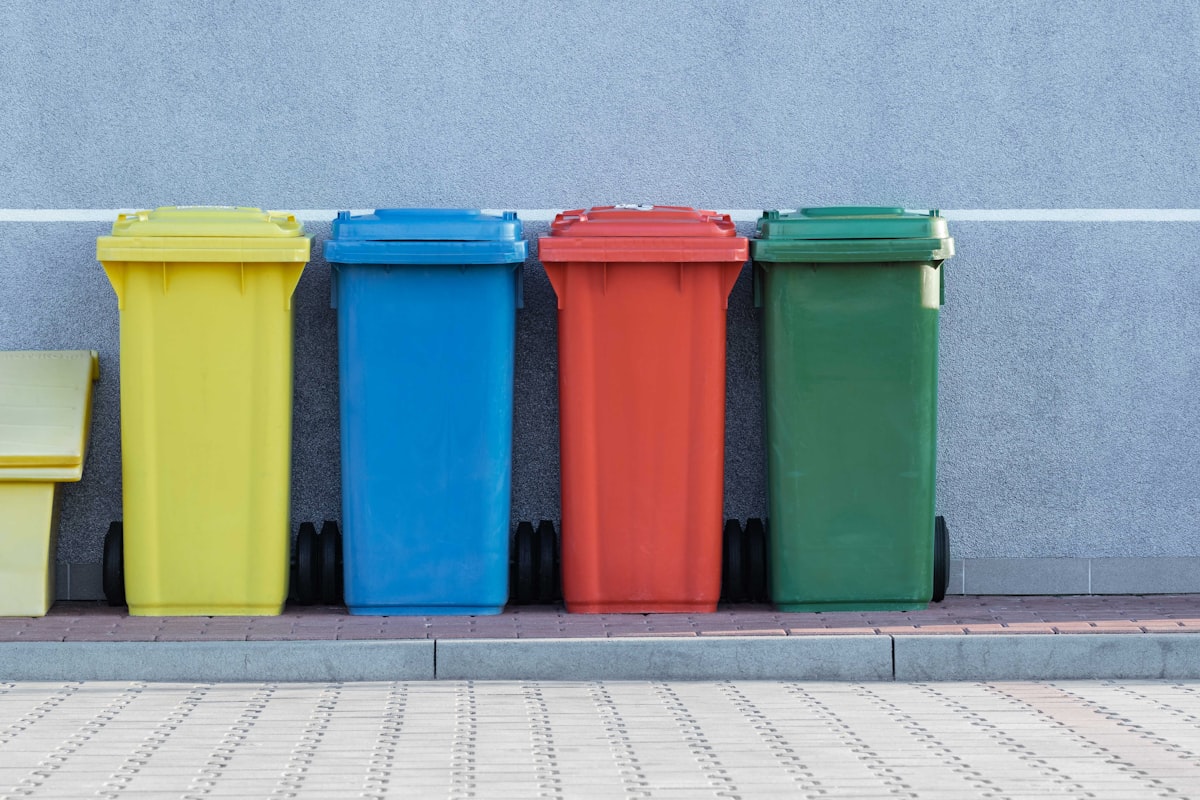 Four color-coded curbside collection bins, yellow, blue, red, and green, lined up along a sidewalk