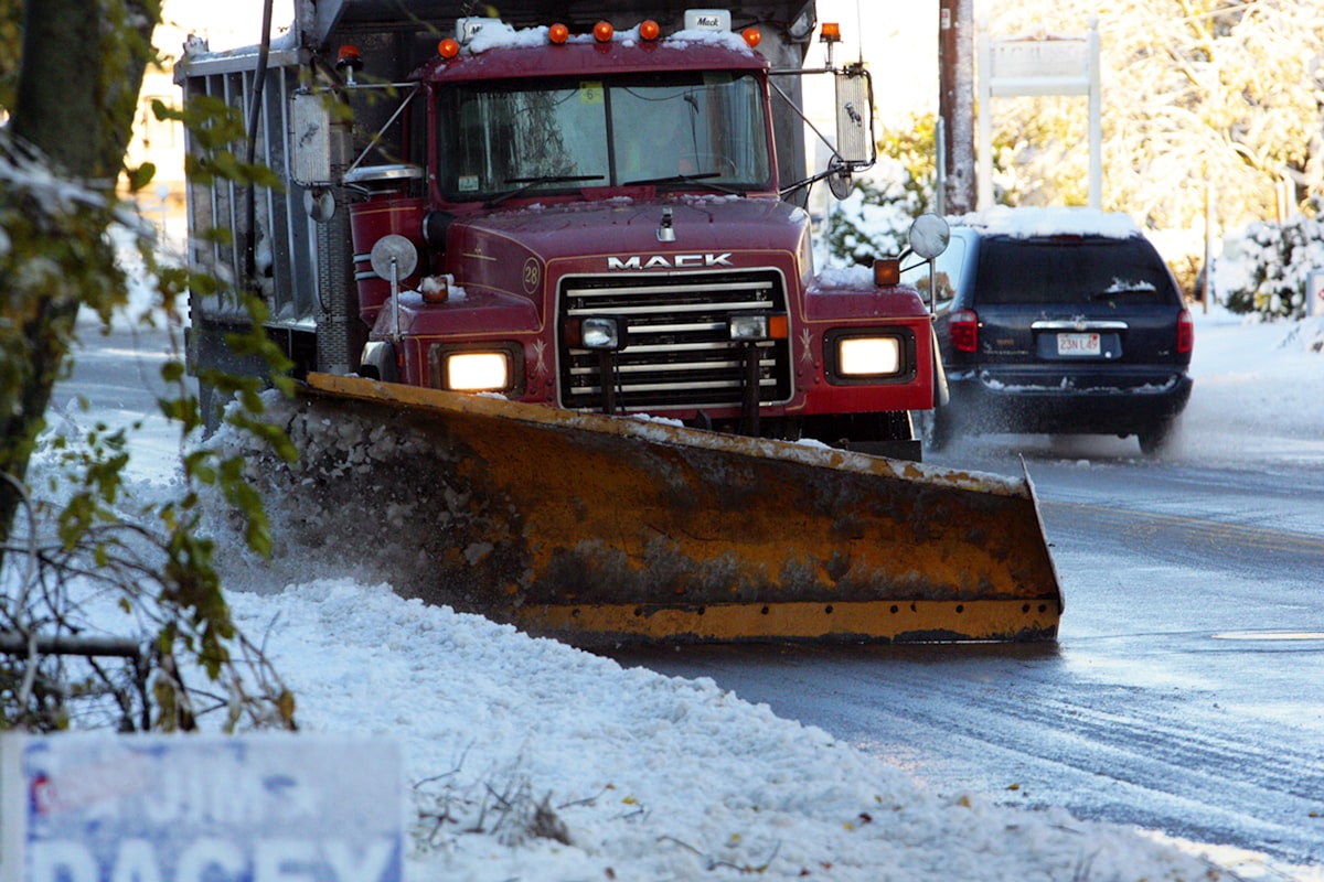A city snow plow truck clearing a residential street after a snowfall, with a car following behind