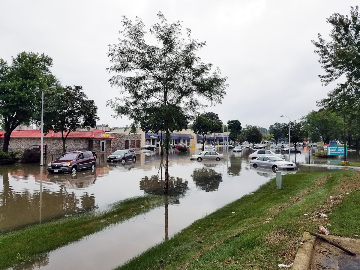 A flooded street after heavy rainfall with cars partially submerged and a storm drain overwhelmed