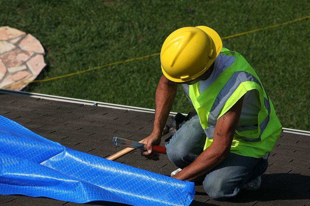 Worker in a hard hat nailing down an emergency tarp on a shingle roof