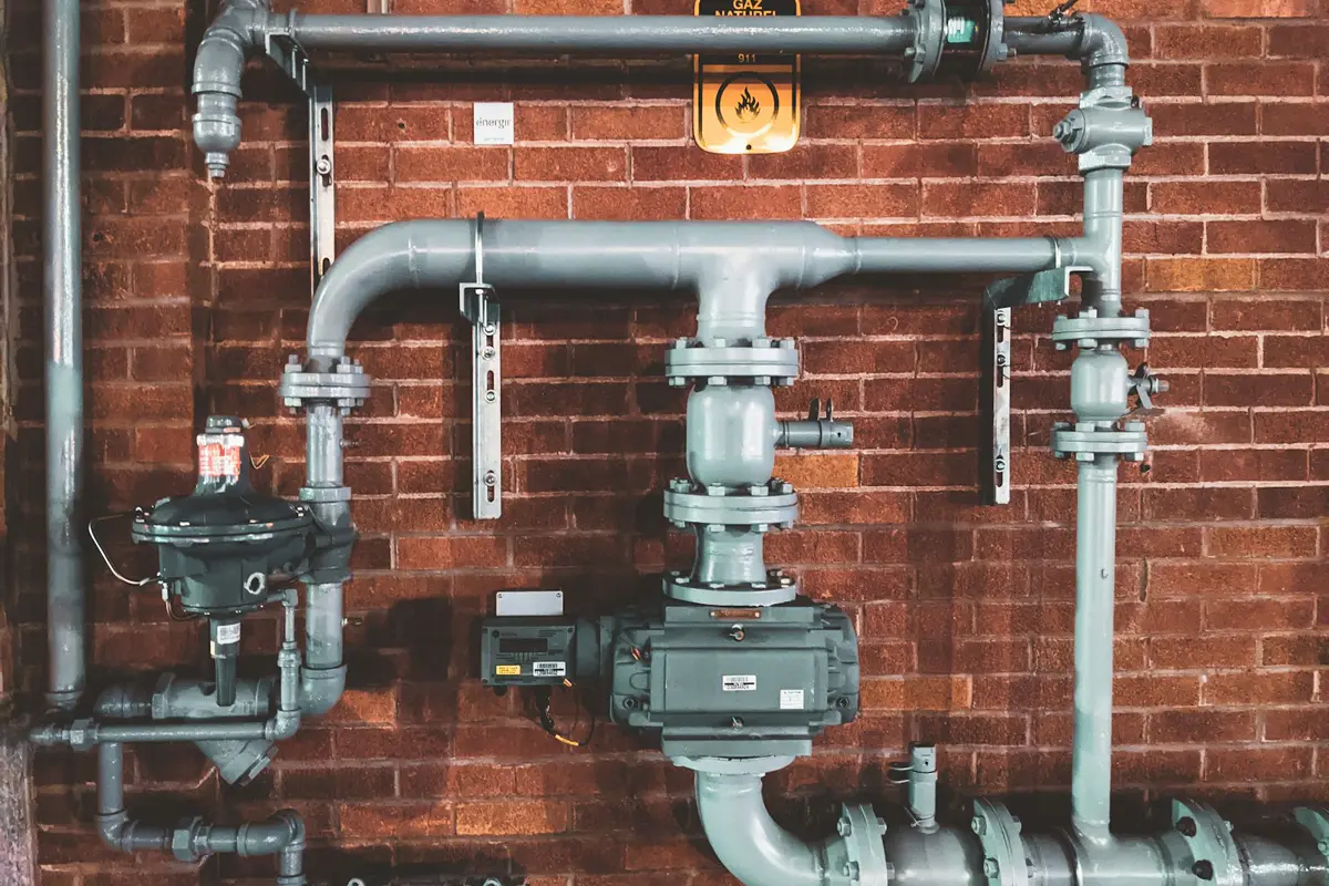 Gray steel water and gas lines with valves and a regulator mounted on a brick wall