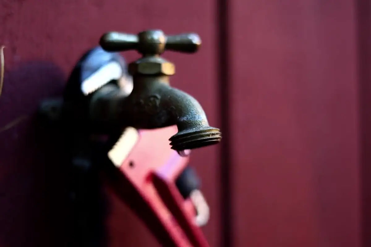 A red pipe wrench gripping an old brass outdoor faucet that is dripping water